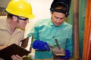 Young welder wearing green overalls with a welding inspector. 