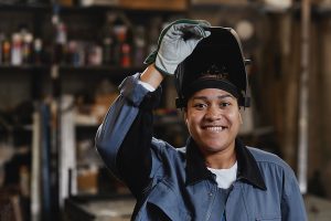 Closeup portrait of a smiling female welder looking at camera while holding up her welding mask. 