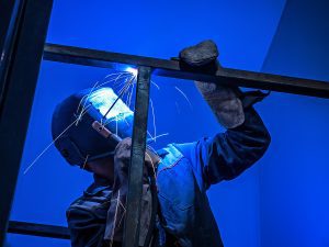 Welder holding a metal beam and performing a task under a blue background. 