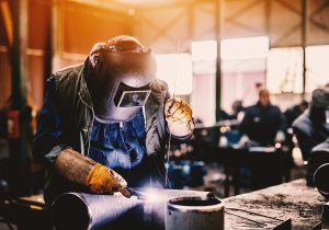 Professional welder in an industrial warehouse setting, welding a metal pipe. 